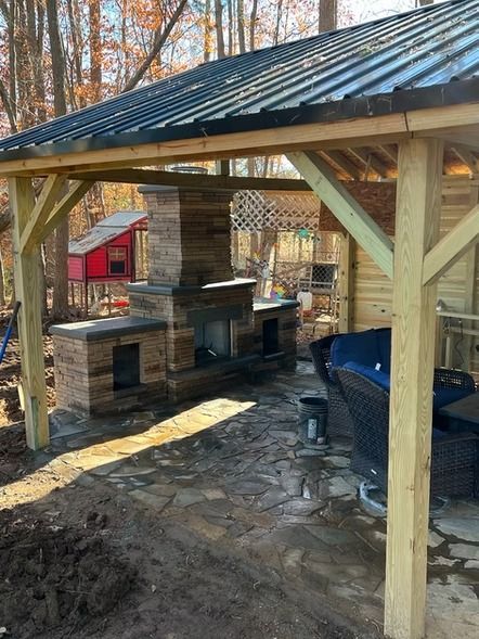 Covered outdoor stone fireplace under a metal roof, with wooden posts and a wooded yard in fall.