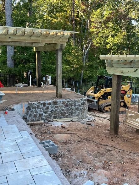 Wooden pergola structures at a construction site, with stone retaining wall, pavers, and a skid steer nearby