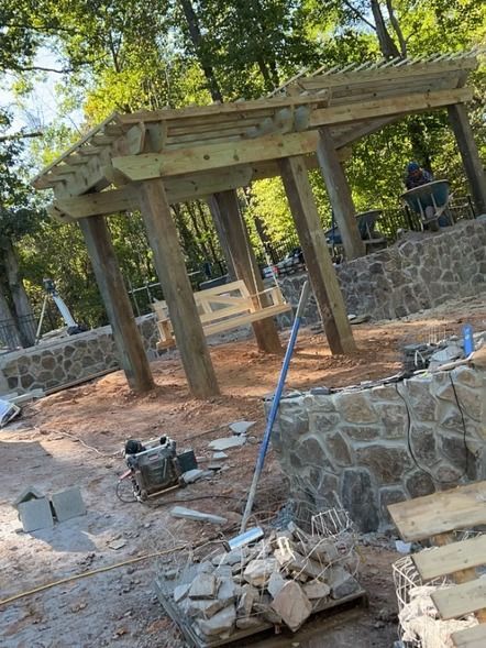Stone pavilion under construction with wooden roof beams, scattered debris, and a blue hose on the ground