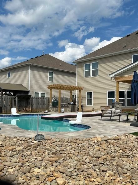 Backyard pool beside beige houses, with patio furniture, pergola, and rock landscaping under a partly cloudy sky