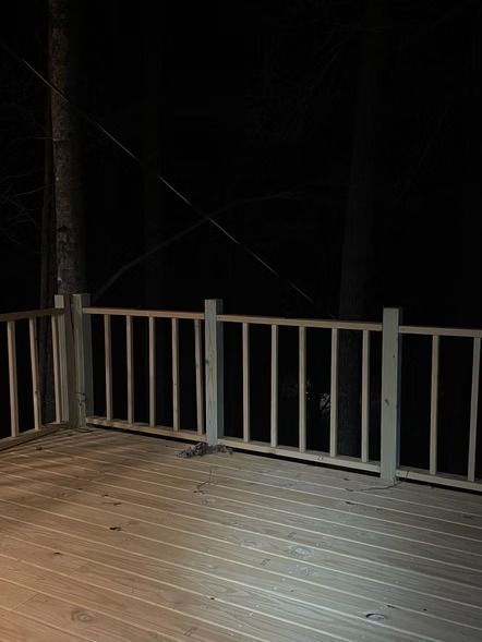 Dim wooden deck with white railing at night, lit by a bright light, overlooking darkness beyond.