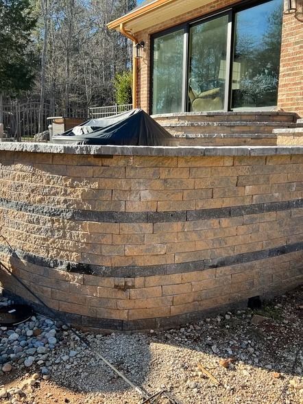 Stone patio wall beside a glass house, with a tarp-covered object and wooded background.