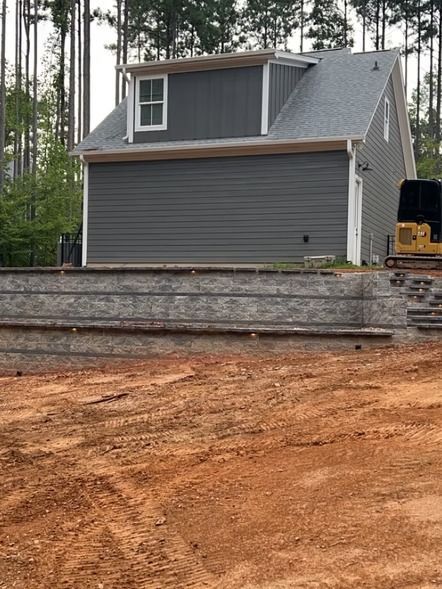 Gray house on a stone foundation at a construction site with red dirt and tall pine trees nearby