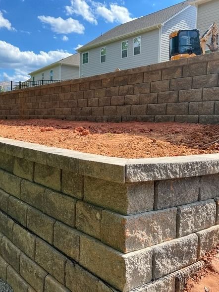 Terraced stone retaining walls with red soil under a blue sky, beside a house on a sloped yard