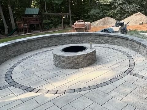 Circular stone fire pit in a paved backyard patio, with trees and construction materials in the background