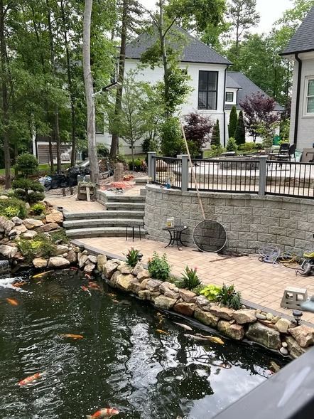Stone patio and retaining wall beside a pond, with steps, railings, and a house in a wooded backyard