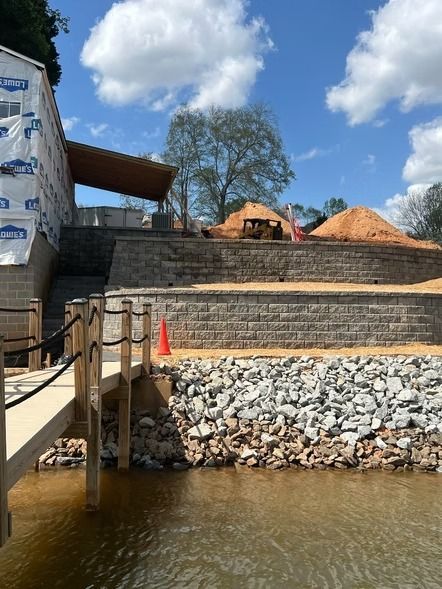 Riverbank construction site with stone retaining wall, rocks, and piles of dirt under a blue sky