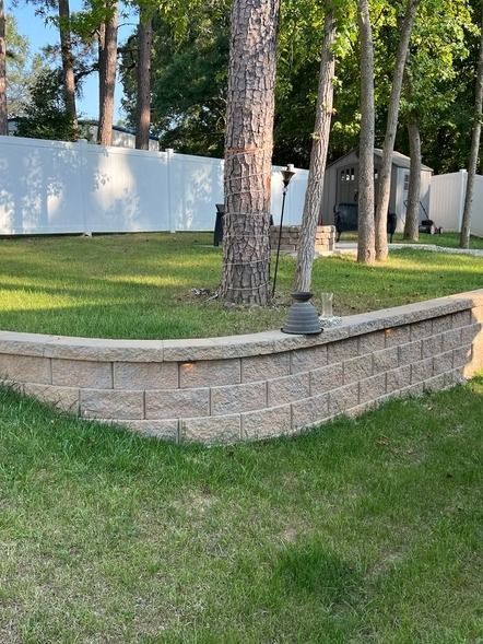 Stone retaining wall beside a grassy yard with pine trees and a white fence in the background