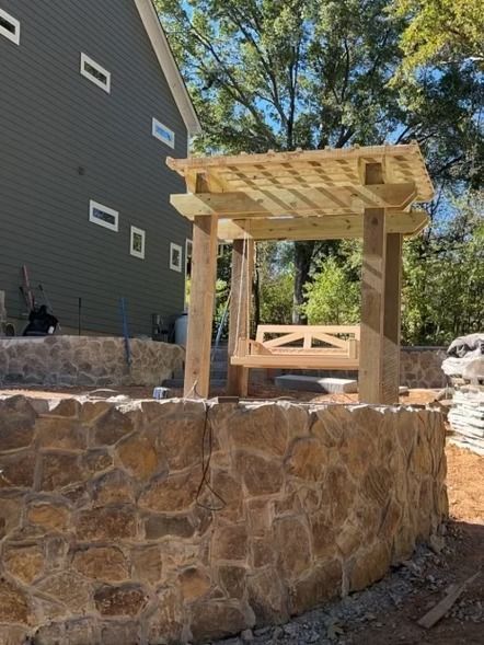 Wooden pergola structure above a stone retaining wall beside a dark house, with trees in the background