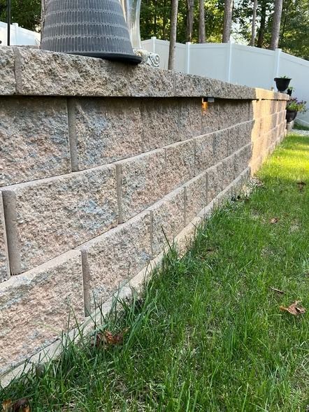 Stone retaining wall beside a grassy lawn, with a black drainpipe and trees in the background