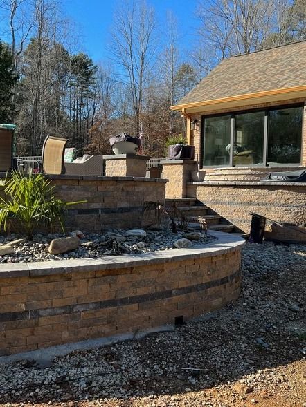Stone patio and brick retaining wall beside a house, with outdoor grill and bare trees in the yard.