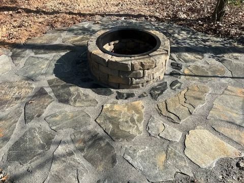 Stone fire pit set in a paved outdoor patio area with a leaf-covered ground in the background
