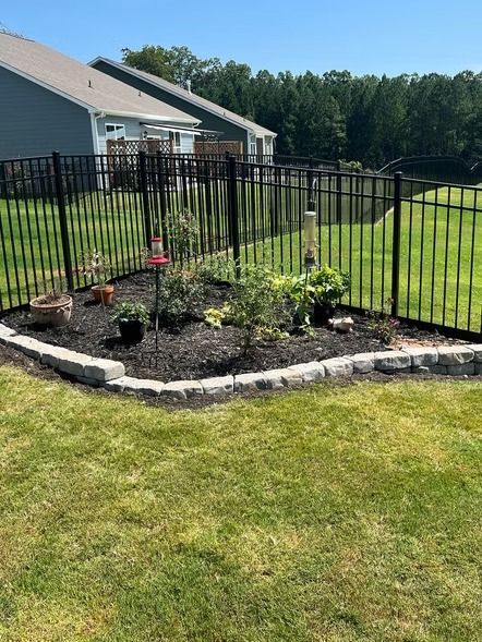Black metal fence around a small landscaped garden bed with flowers beside a house and lawn