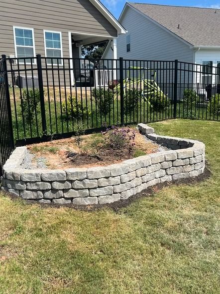 Small raised stone garden bed with sparse plants in a front yard beside a black metal fence.