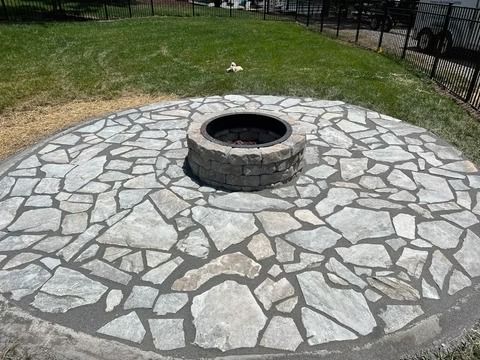 Stone fire pit centered in a circular paved patio with grass and a fence in the background