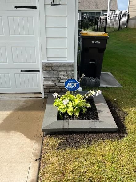 Front porch with a black planter of yellow flowers beside a garage and driveway, next to an ADT sign and trash bin
