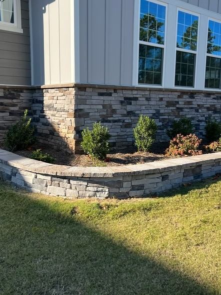 Stone retaining wall with small shrubs in front of a house with light siding and windows.