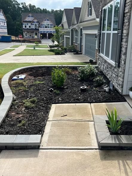 Residential walkway beside gray houses with landscaped garden beds and concrete path