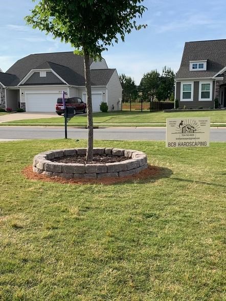Front yard with a young tree in a stone ring and a neighborhood sign near suburban houses.
