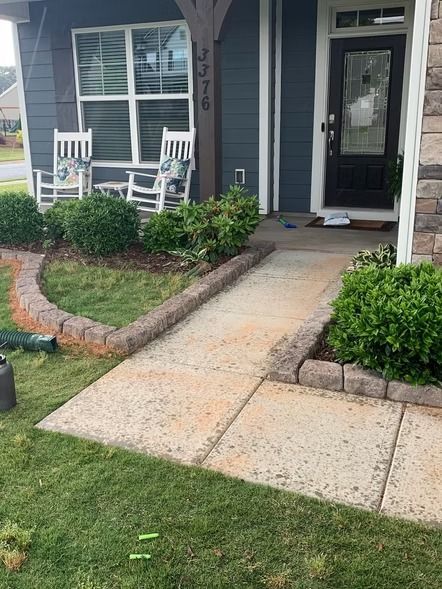 Front porch with concrete walkway, white rocking chairs, dark exterior door, and neatly landscaped shrubs.
