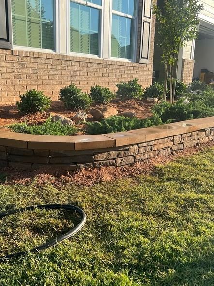 Raised brick garden bed with shrubs and mulch beside a house, with a garden hose on the lawn.