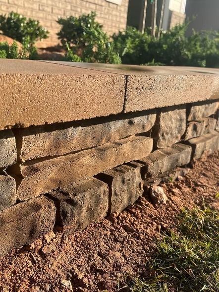 Close-up of a stacked stone retaining wall with a tan capstone, beside a dirt and grass yard.