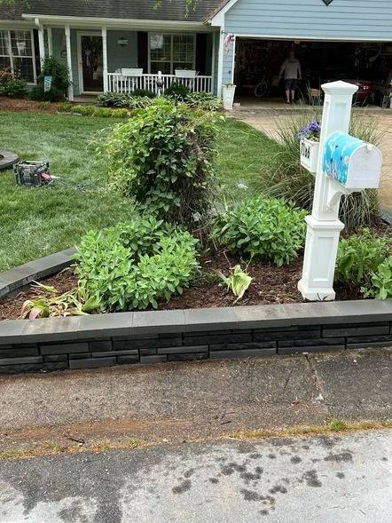 Front yard garden bed beside a driveway, with a white mailbox and a porch in the background