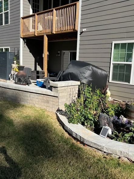 Backyard patio with grill, covered furniture, wooden balcony, stone retaining wall, and flower bed