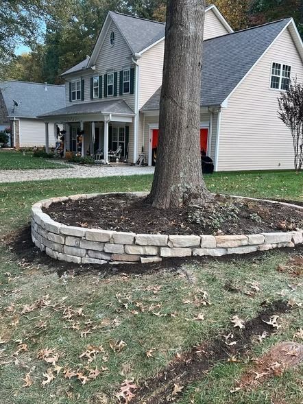 Stone-edged tree ring in a front yard beside a gray two-story house with a porch