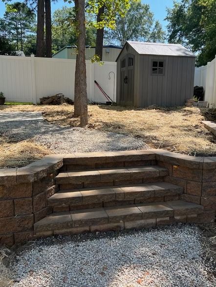 Backyard steps leading to a gravel patio, with a tree and gray storage shed by a white fence.