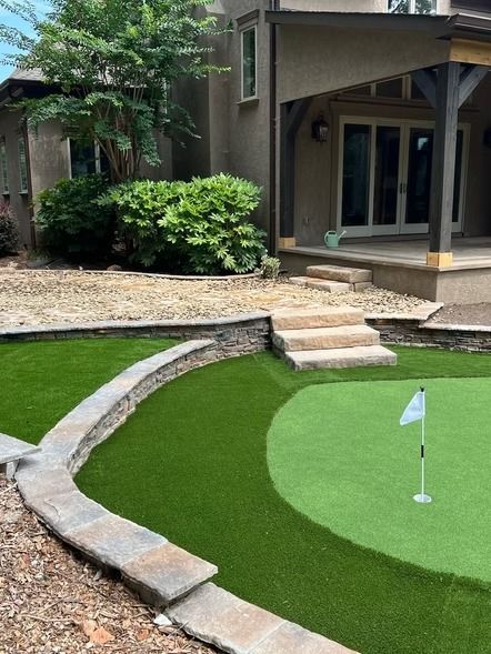Small backyard putting green with a flag, stone border, and steps beside a patio.