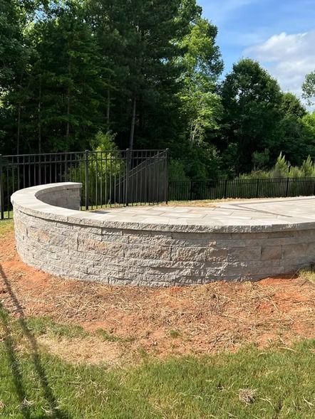 Curved stone retaining wall beside a black metal gate in a grassy yard with trees and blue sky