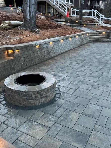 Stone plaza with a round fire pit and low retaining wall lined with small lights near stairs and trees