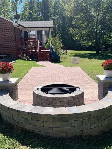 Backyard patio with a circular stone fire pit, brick walkway, and lawn bordered by trees