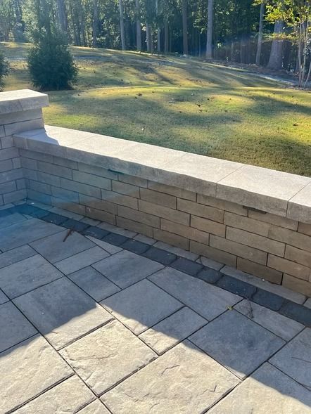 Stone patio beside a low brick wall overlooking a sunlit grassy yard with trees.