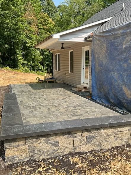 Stone patio beside a beige house with a blue tarp and wooded yard