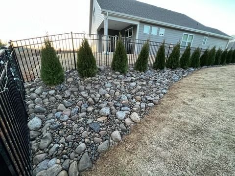 Rock garden with river stones and evergreen shrubs beside a house and lawn, bordered by a black fence