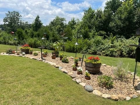 Curved garden bed with potted flowers, stones, and shrubs beside a lawn under a partly cloudy sky
