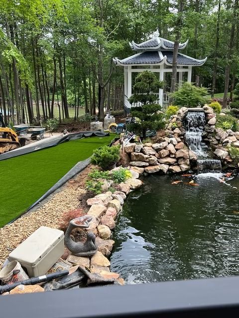 Backyard pond with waterfall, stone edging, and a white gazebo amid trees