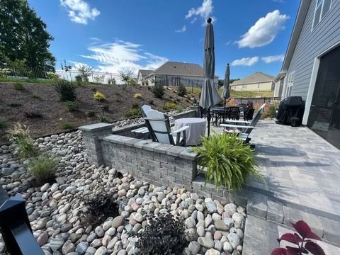 Backyard stone patio with outdoor dining chairs, umbrella, landscaped rocks, and green hillside under blue sky