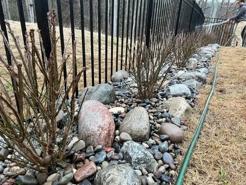 Rock garden bed with river stones, bare shrubs, and a black metal fence along a grassy path.