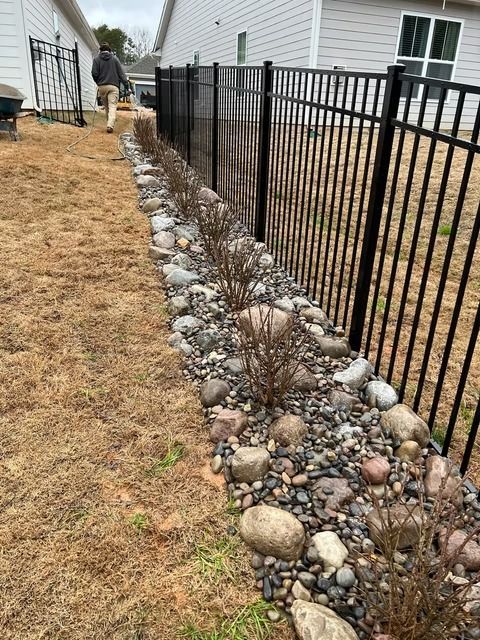 Rock-lined garden bed with small shrubs beside a black metal fence and white house