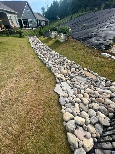 Stone drainage swale lined with rounded rocks beside a suburban lawn and house.