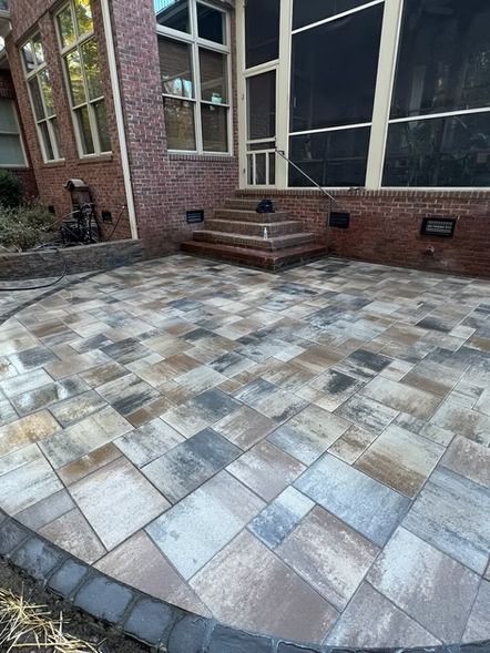 Brick patio with gray and tan pavers beside a house, with steps leading to a glass door.