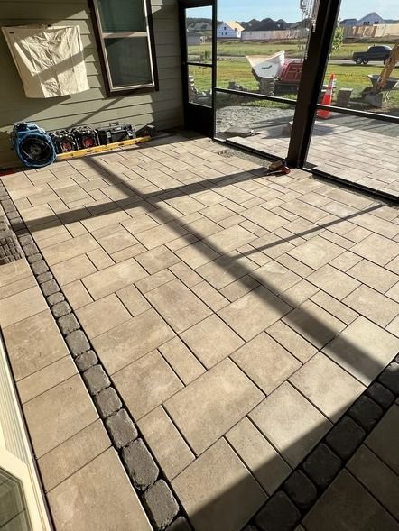 Outdoor patio with light paver tiles and dark border, viewed through a screened enclosure.
