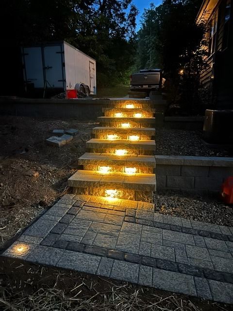 Lit stone steps at dusk lead up a driveway beside a house and parked trailer.