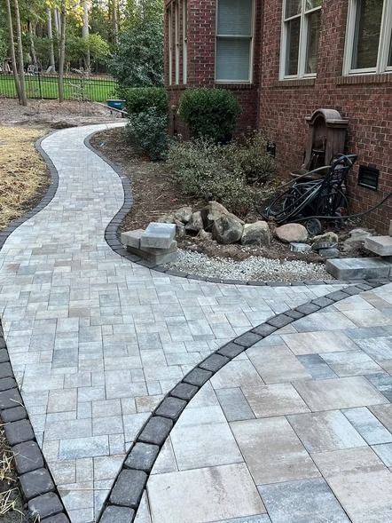 Curved light-gray paver walkway beside a brick house and landscaped garden beds