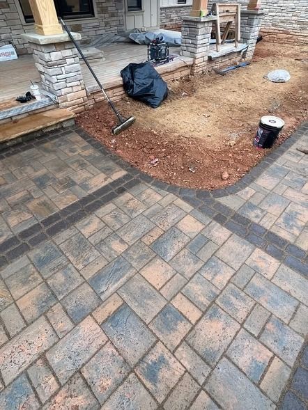 Brick patio beside a stone porch with a dirt patch, shovel, black bag, and bucket.