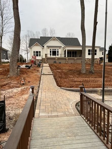 Stone walkway leading to a large white house under construction, framed by bare trees and unfinished landscaping