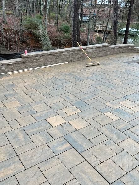 Stone-paved patio with a broom leaning against a low retaining wall overlooking trees and a stream